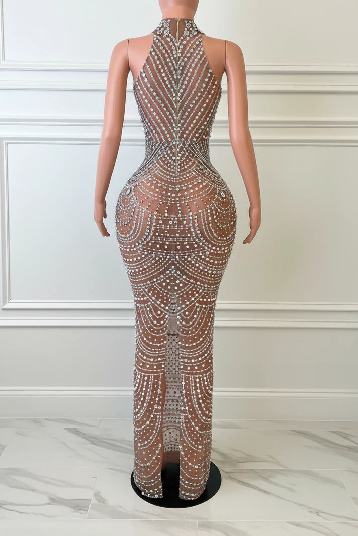ManneqYembi Fashion silver rhinestone maxi dress with high neck choker collar, plunging cutout bodice, draped chest chains, and sheer nude illusion meshuin wearing a sparkling jumpsuit in front of a decorative mirror.